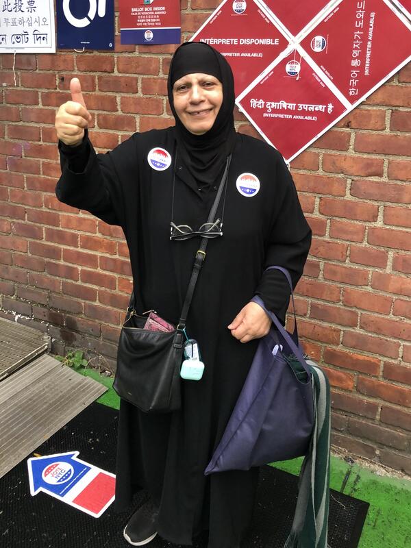 Maha outside of her polling place after she votes for the first time Maha stands in front of a brick building giving a thumbs up and wearing two stickers that say 'I voted early." There are signs behind her in various languages with directions for voting.