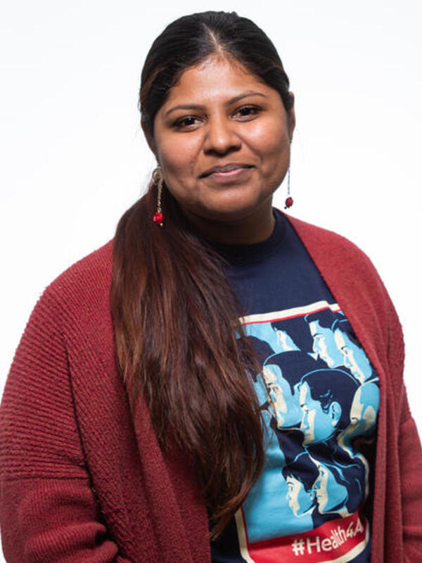 Lupe, a DACA recipient and youth leader Lupe smiles and looks at the camera while wearing a blue shirt that says #Health4All and a red cardigan.