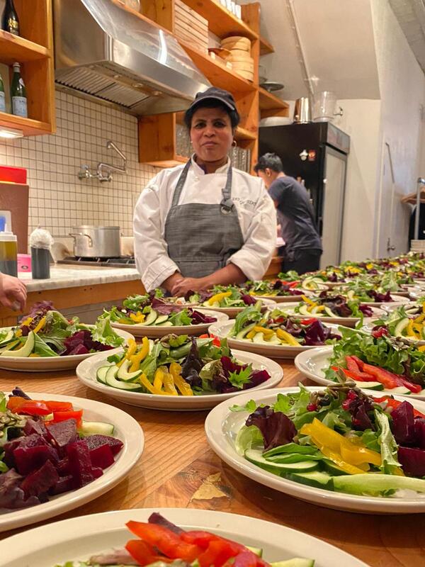 Wearing an apron, Chef Shanthini stands behind a table in a kitchen with many plates of a salad. 