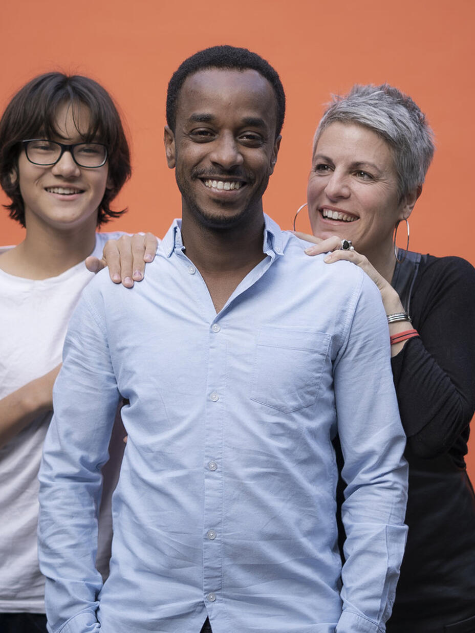 Eine Frau und zwei junge Männer stehen vor einer orange farbenden Wand