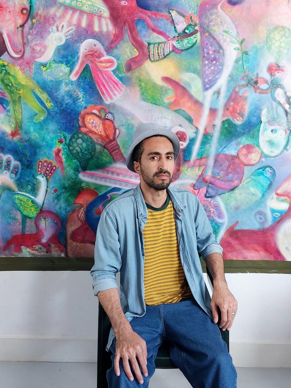 Nima Javan sits in front of his artwork, which is a large painting of colorful sea creatures.