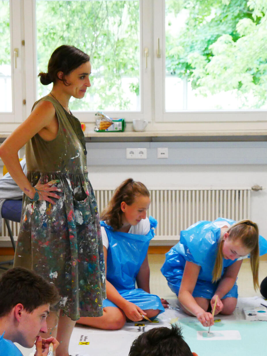 Diala stands with her hands on her hips in a room with children who are painting 