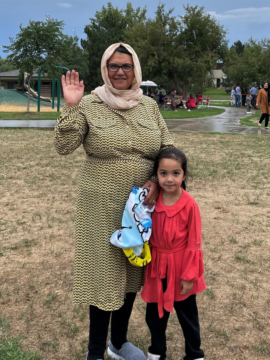 A woman with a hijab waving and smiling with a little girl in pink at a park.