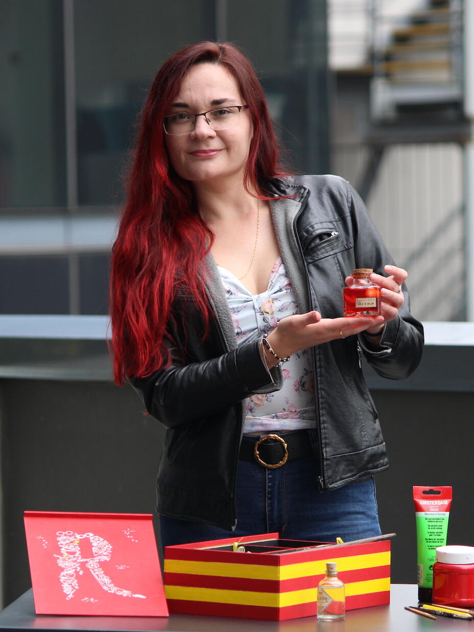 Una mujer sostiene un frasco de líquido rojo.  Frente a ella hay una mesa sobre la que se exponen otros objetos coloridos.