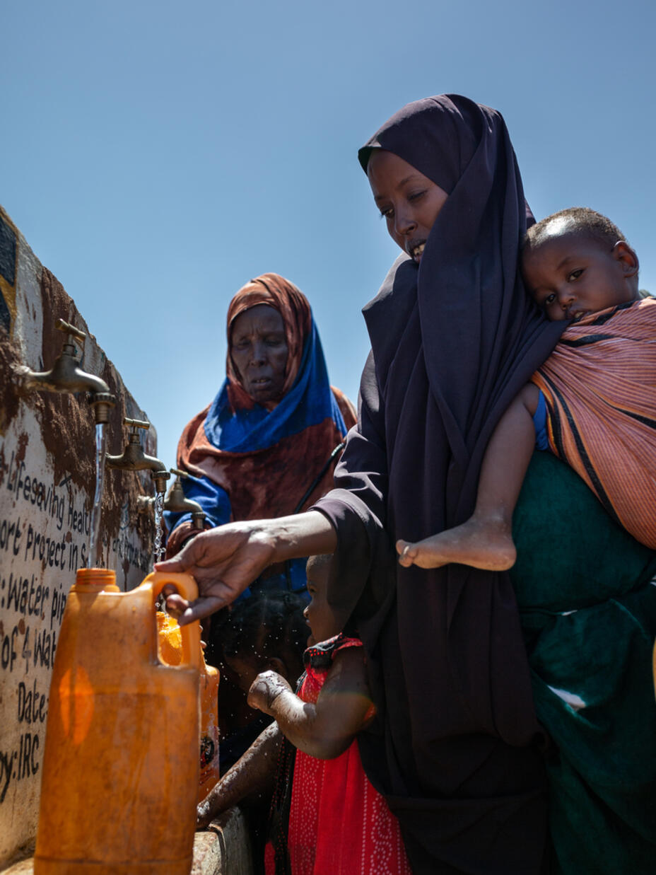 Two women fill up water containers. Ones holds a baby on her hip as she does so.