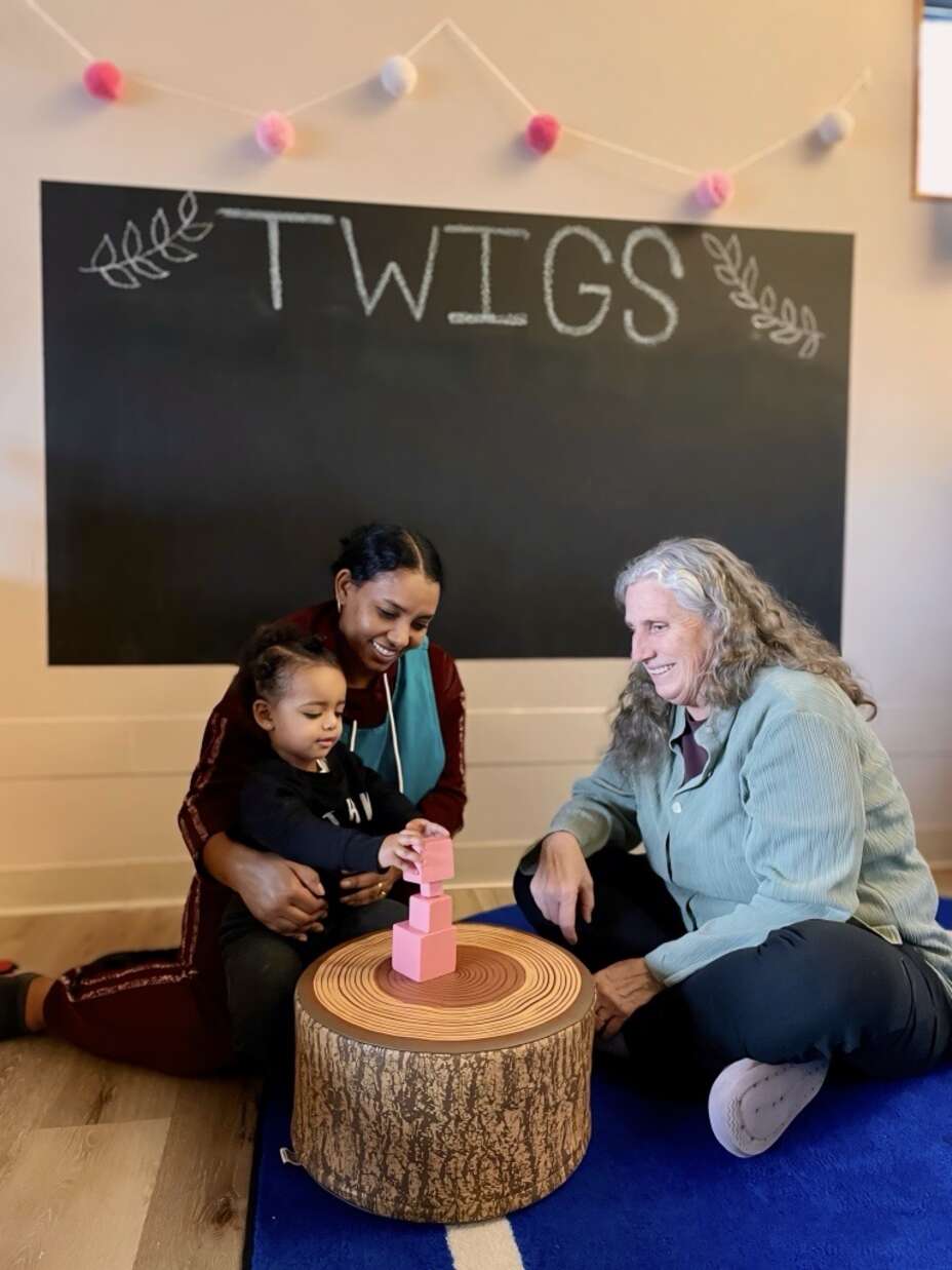 A young woman with a child on her lap sits across from an older woman. They are both smiling and looking down at the child who is playing with pink blocks.