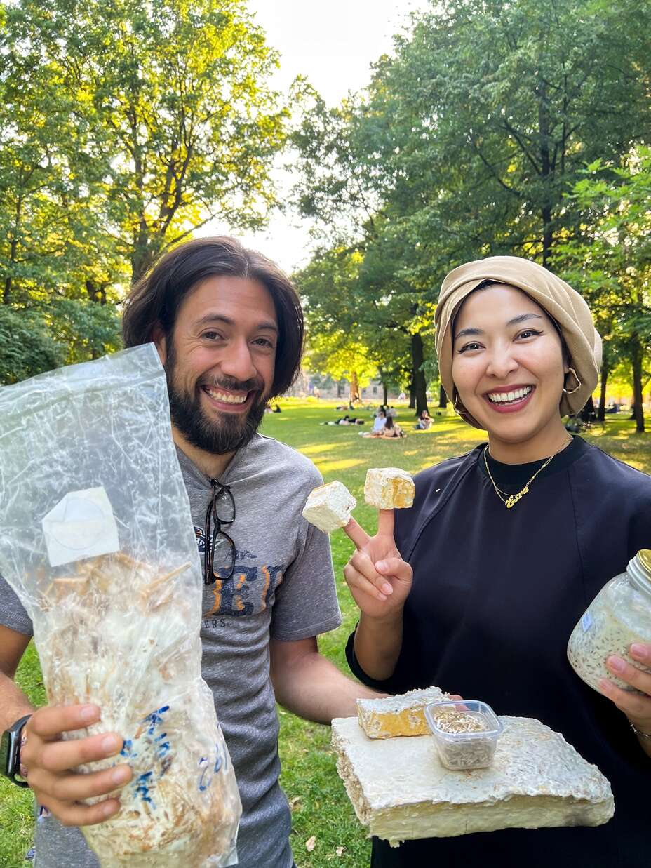 Ein Mann und eine Frau stehen in einem sonnigen Park und lächeln in die Kamera. Der Mann hält eine Plastiktüte mit fermentierten Produkten, während die Frau eine Platte mit weiteren fermentierten Lebensmitteln und ein Glas mit fermentierten Samen präsentiert.