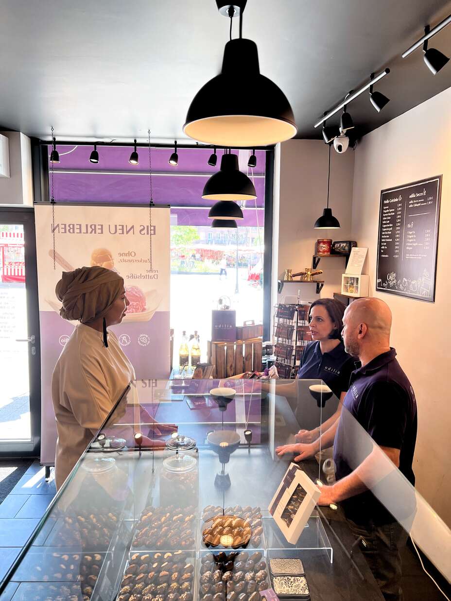 Esra Karakaya steht an einer Theke in einer Schokoladenboutique und spricht mit zwei Mitarbeitern. Die Boutique ist modern eingerichtet, mit einer Auswahl an Schokoladenprodukten in einer Glasvitrine und Werbebannern im Hintergrund, die nach außen zu einer belebten Straße zeigen.