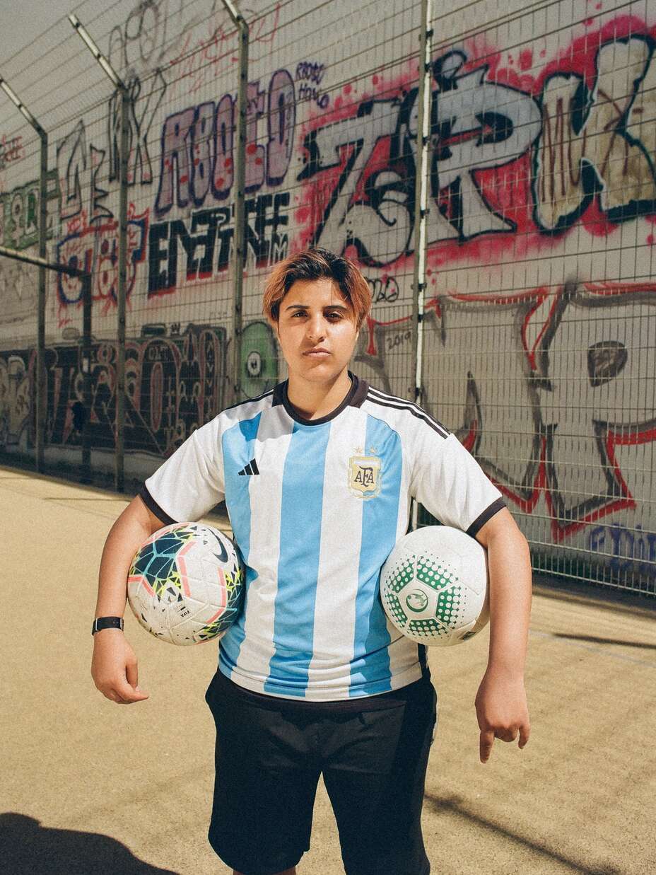 Eine Fußballspielerin in einem argentinischen Trikot hält zwei Fußbälle vor einer mit Graffiti bedeckten Wand.