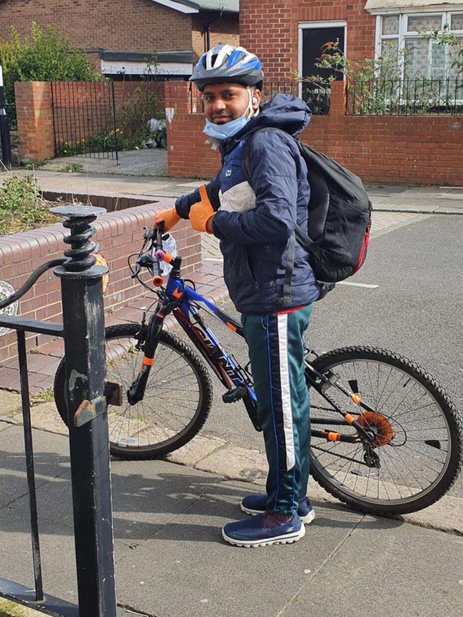 Md Mominul Hamid stands next to his bike and gives a thumbs up. He is wearing a blue helmet and orange gloves and a black backpack. 