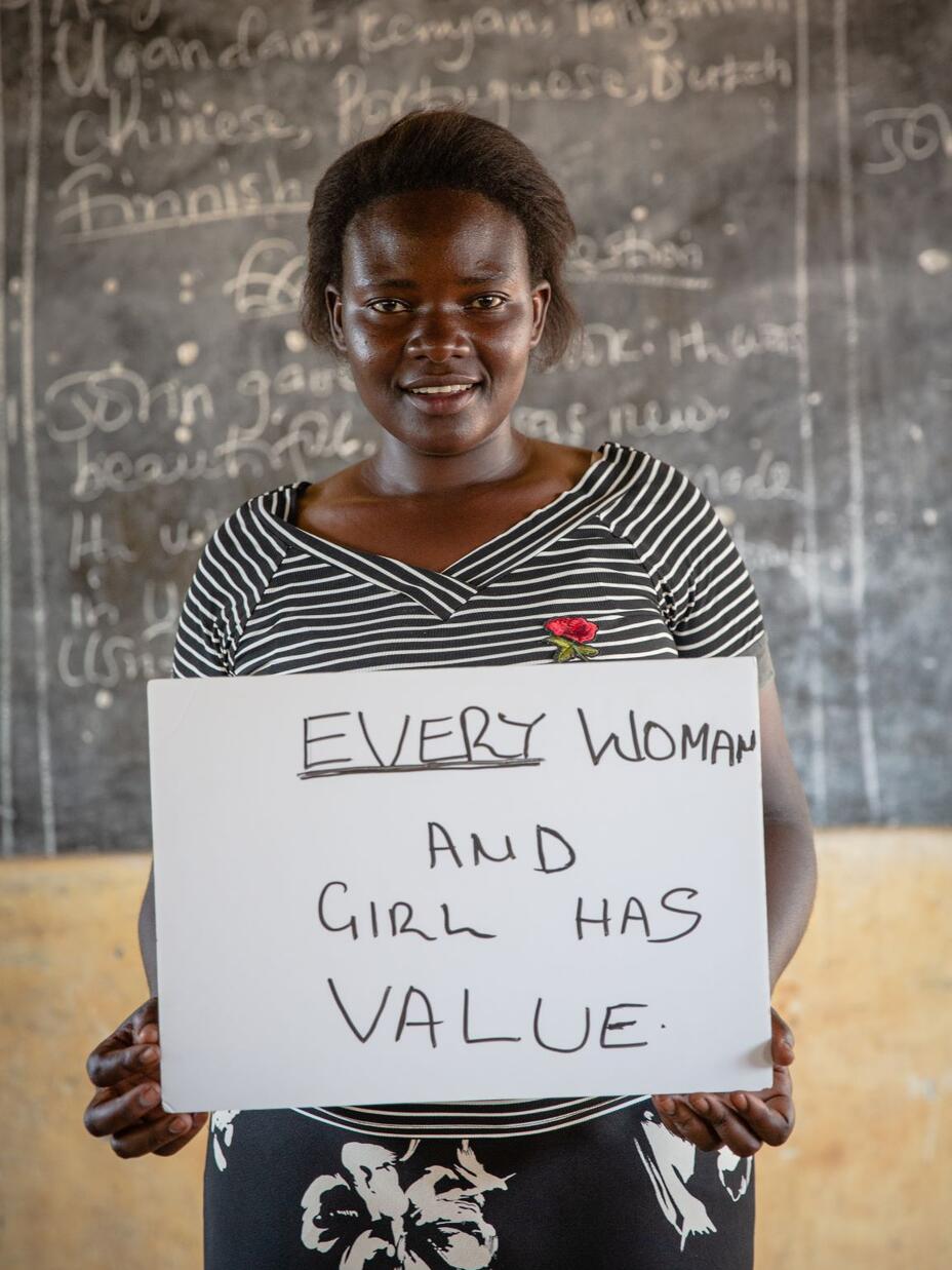 Jackie Letaru, wearing a black and white striped shirt and standing in front of a chalkboard, holds a sign that says "Every woman and girl has value." 