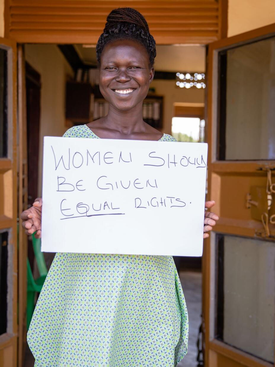 Grace Muuduru, smiling and looking at the camera, holds a sign that says "Women should be given equal rights"