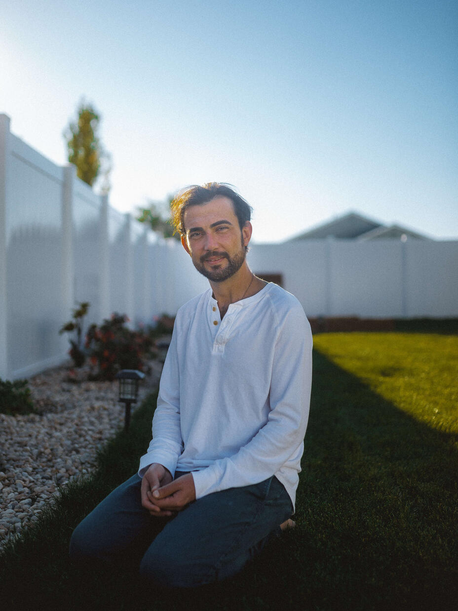 Shadi, wearing a white botton-down shirt, sits in his backyard in front of his white fence near his garden. 