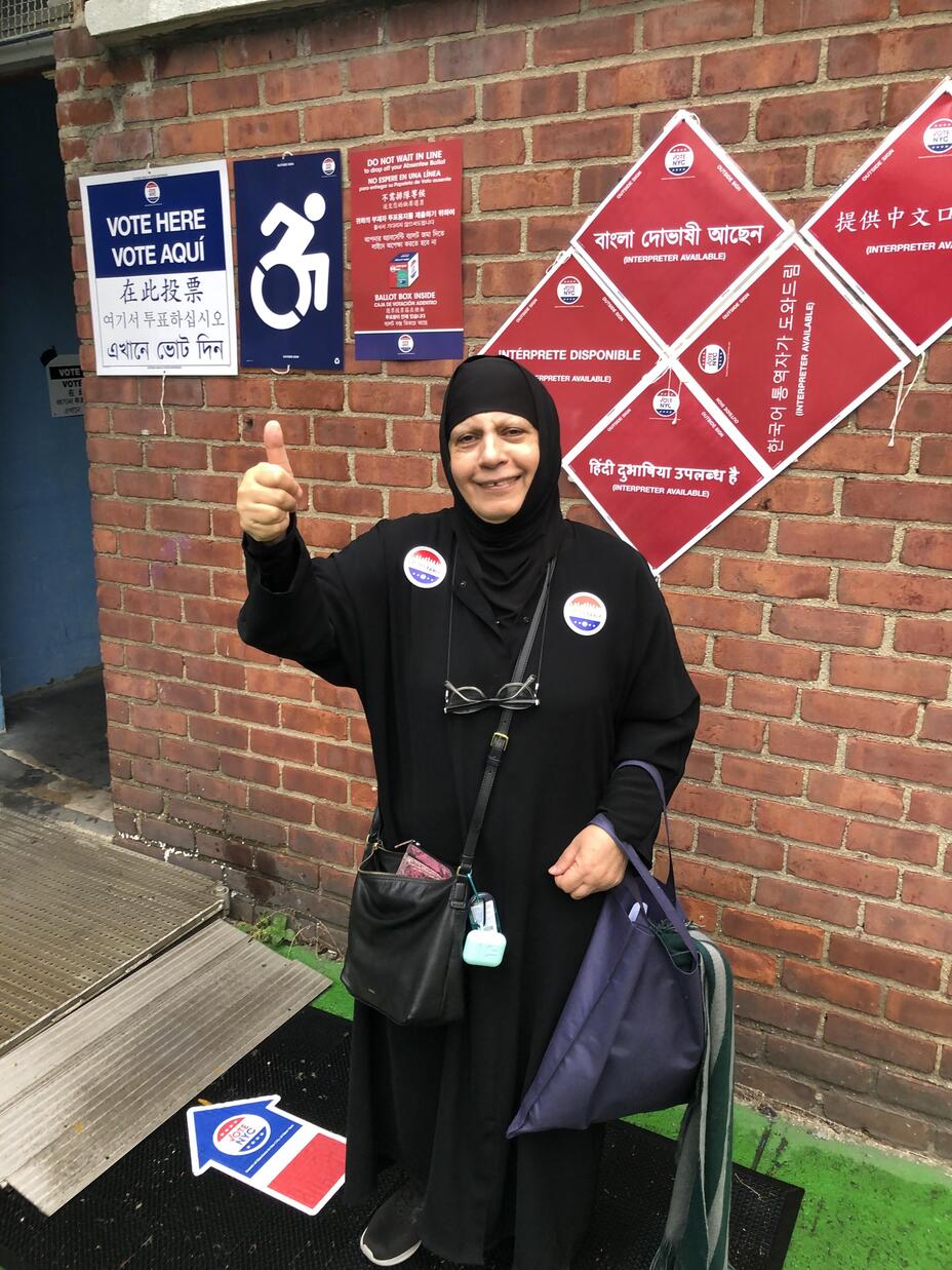 Maha stands in front of a brick building giving a thumbs up and wearing two stickers that say 'I voted early." There are signs behind her in various languages with directions for voting.