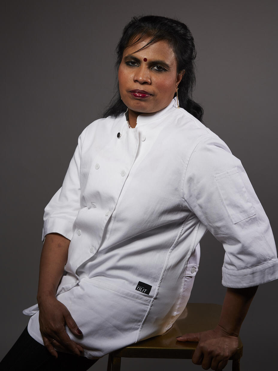 Chef Shanthini, wearing her chefs coat, sits on a stool and looks at the camera. 