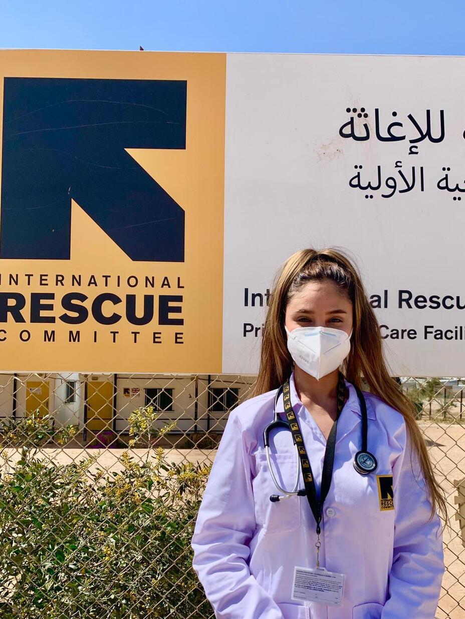 Wearing a mask, Dr. Rose Al-Nsour stands outside in front of an IRC logo.