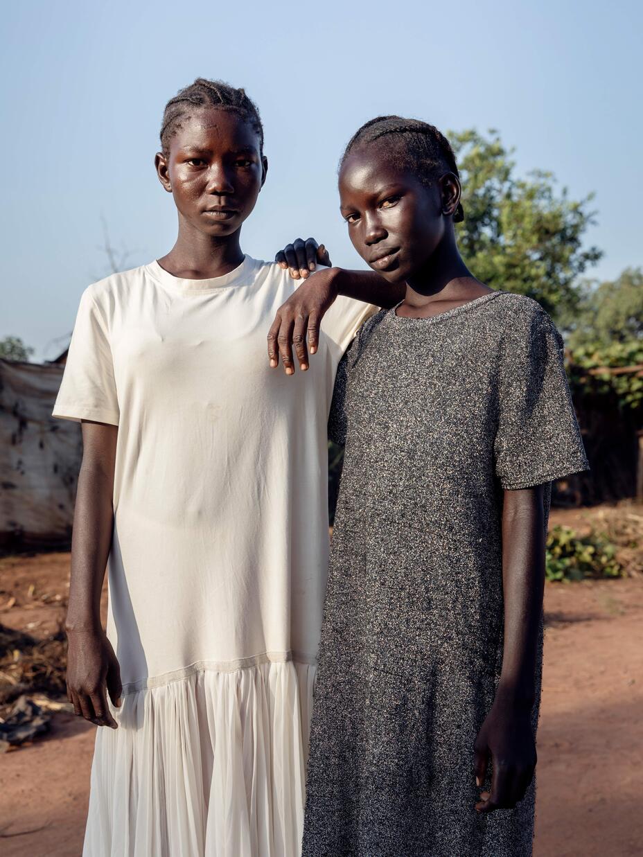 Two girls stand, with one leaning on the other and both looking at the camera. 