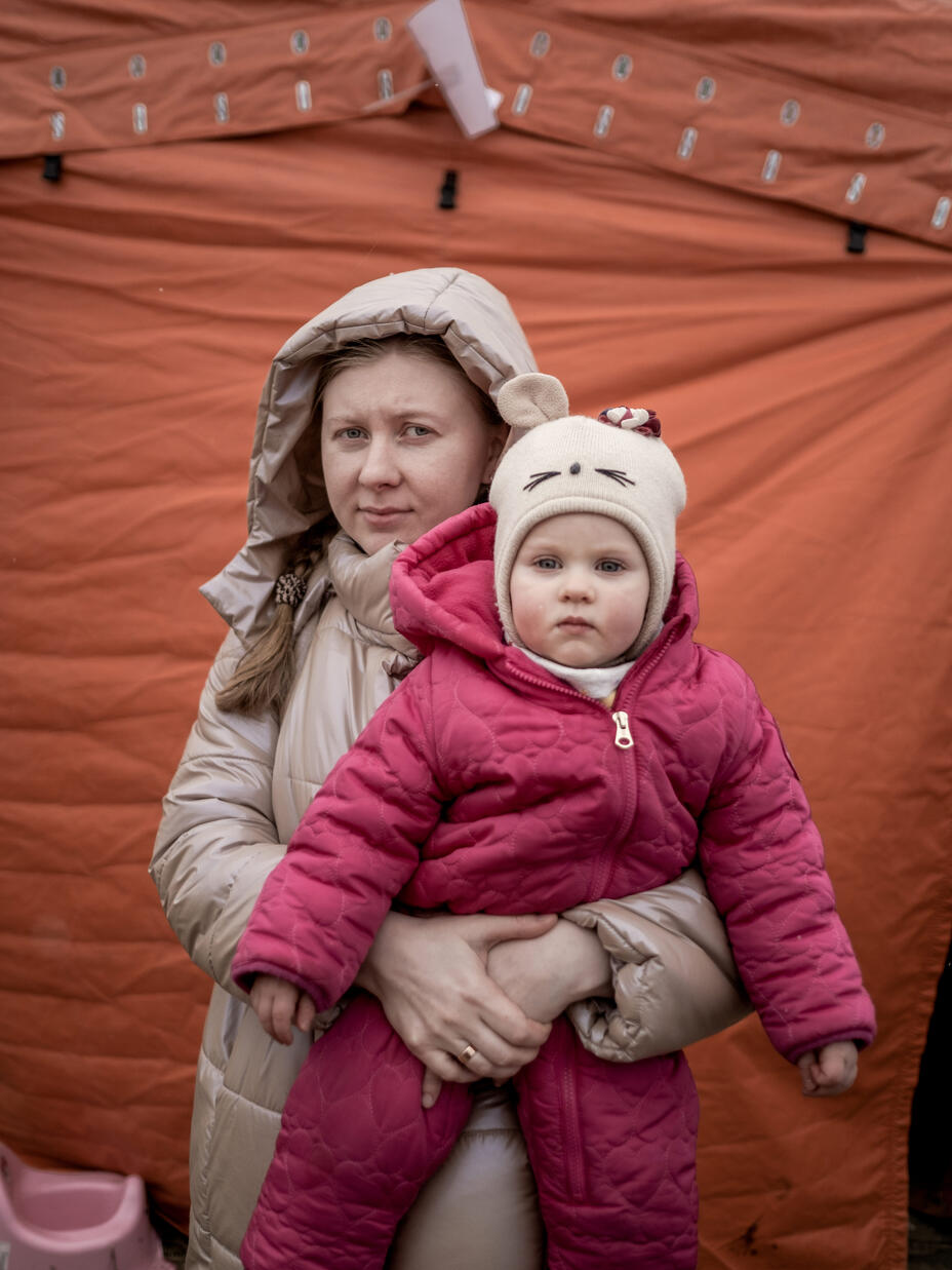 Iryna, a refugee from Ukraine, holding her daughter. They are standing in front of a tent and wearing all winter clothes. 