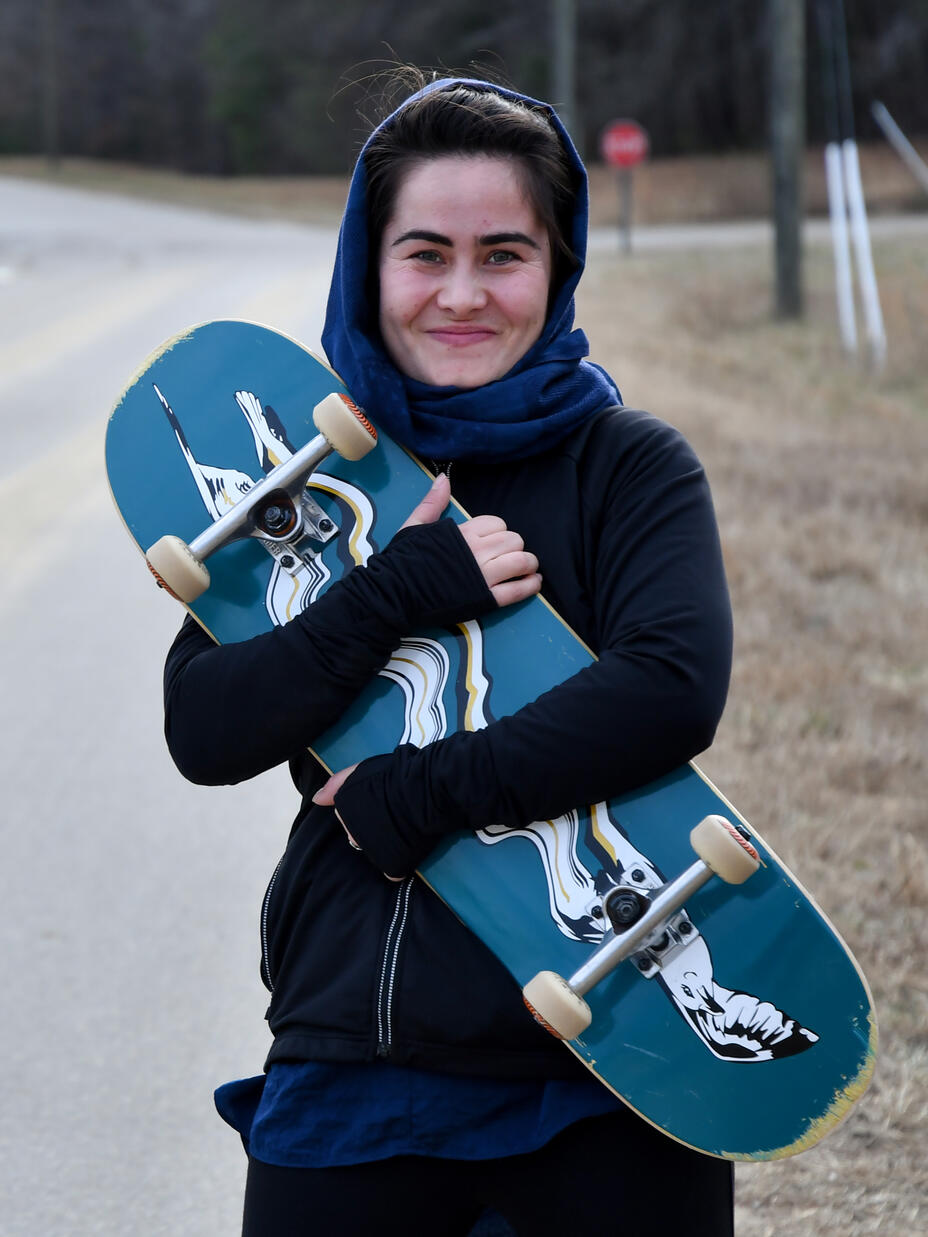 Belqisa holds her skateboard to her chest and smiles looking at the camera 