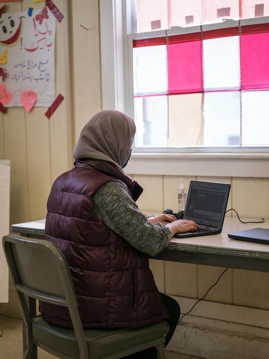 A young woman, with her back to the camera, sits at a desk and types on her laptop.