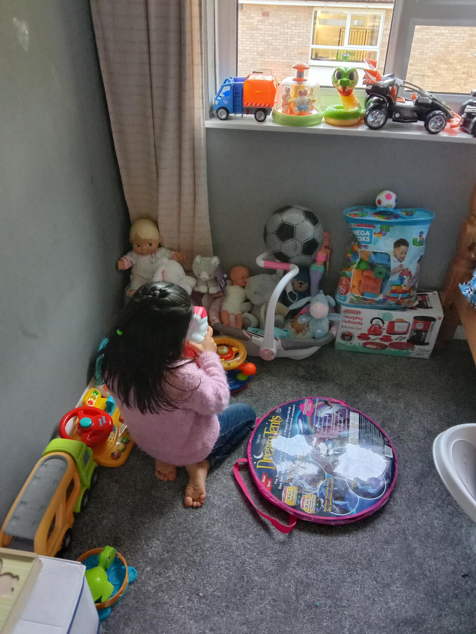In a carpeted room, Sahir's daughters stands among her toys 