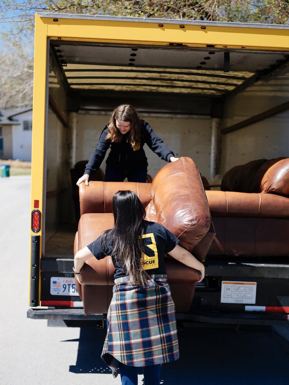 Two women moving a couch out of a moving truck parked curbside.