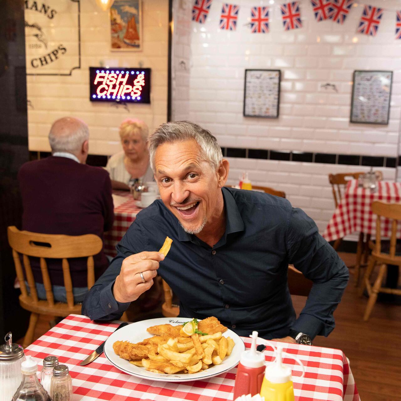 Gary Lineker sitzt an einem Tisch und isst Fish & Chips.