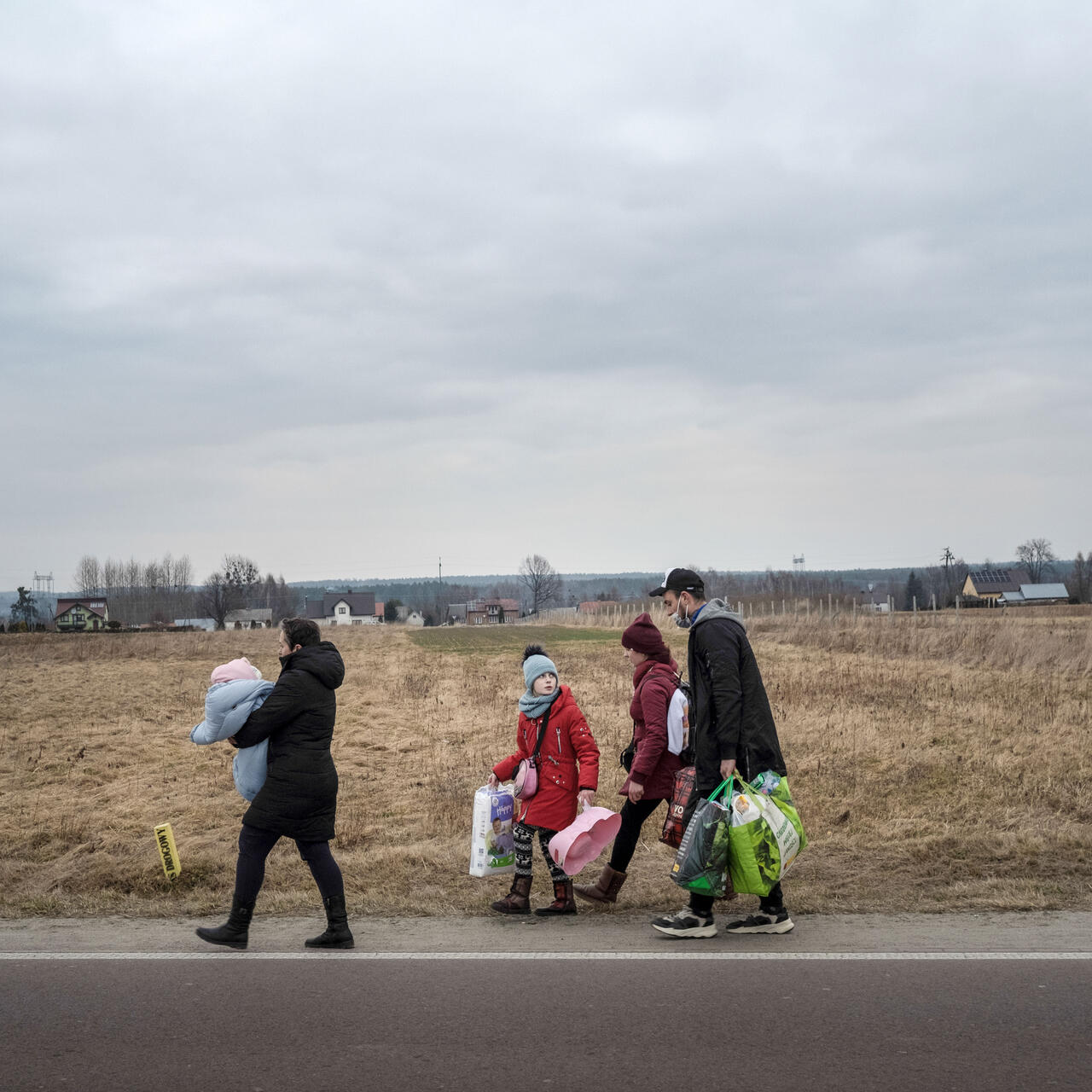 Eine Familie mit drei Kindern läuft zu Fuß mit vielen Taschen und Tüten eine Landstraße lang