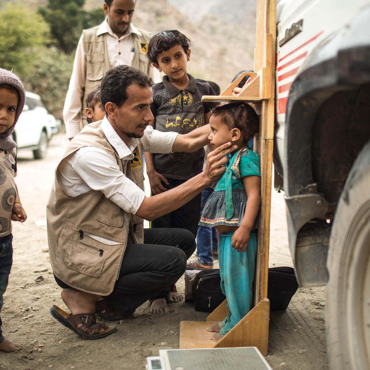 IRC 이동진료소의한남성의료진이예멘의외딴산지마을에서영양실조 징후를보이는아동을진찰하고, 다른아동들이그장면을지켜보고있습니다.