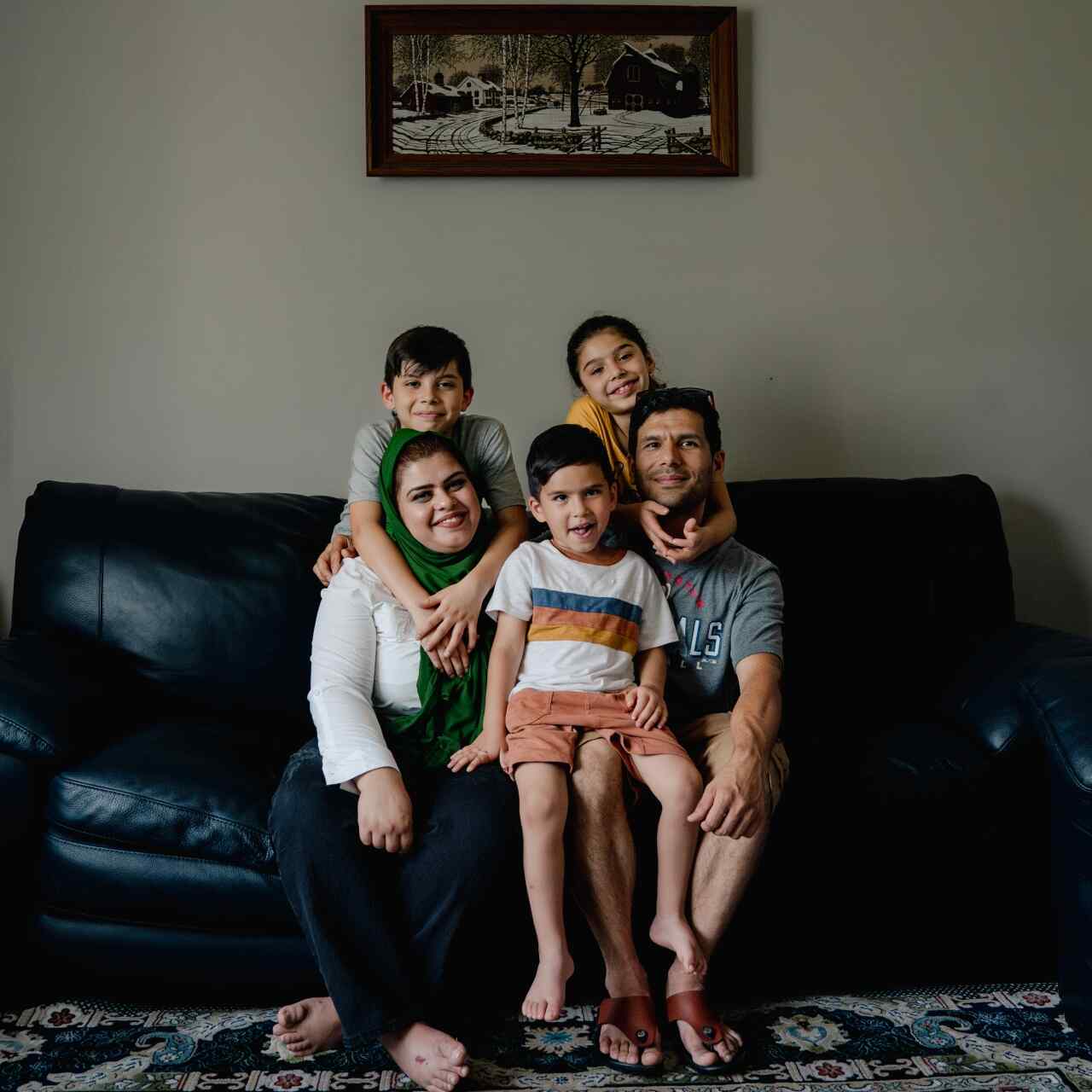 Two parents and three children pose for a portrait together on a black couch. The family was resettled after fleeing Afghanistan as refugees.