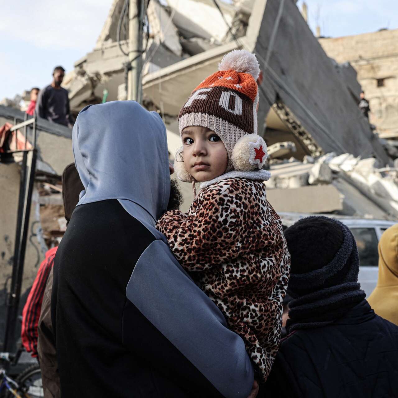 A young girl looks away from a destroyed building in Palestine while in her father's arms.