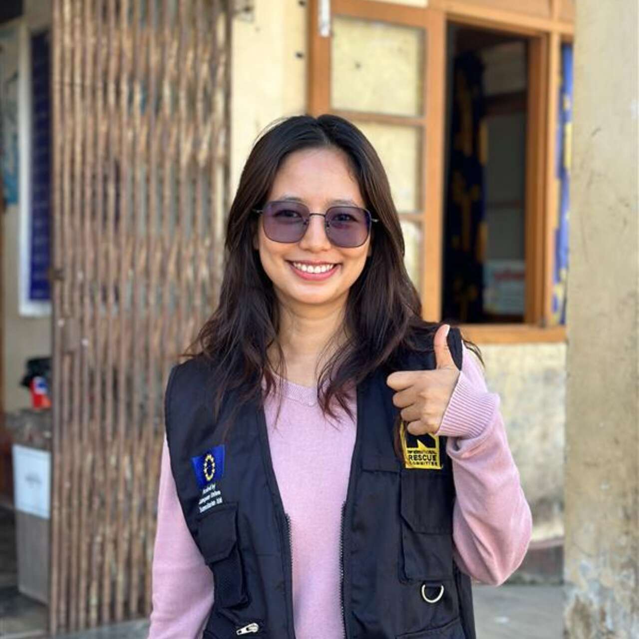 A young woman with long brown hair and sunglasses is looking at the camera and doing a thumbs up with her left hand