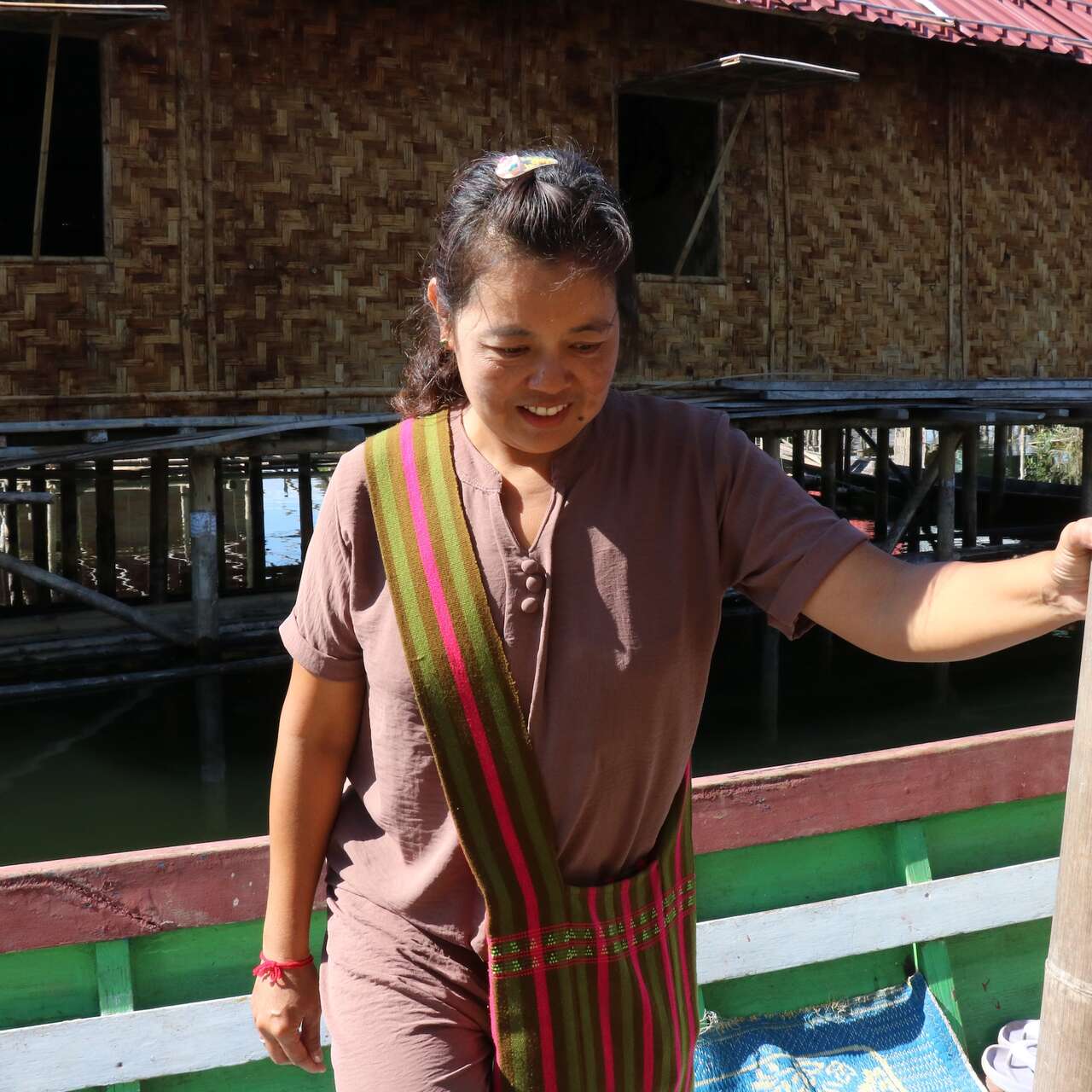 A woman on a boat wearing a brown shirt and carrying a pink and green crossbody bag looks down and smiles