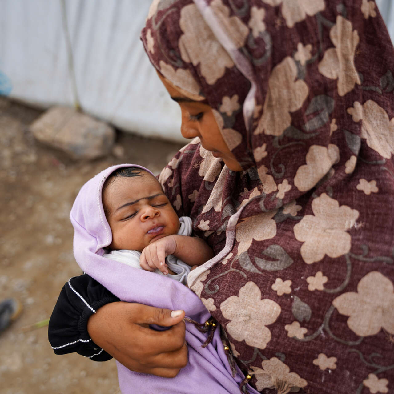 En mamma står utanför ett tält och håller i sin bebis.
