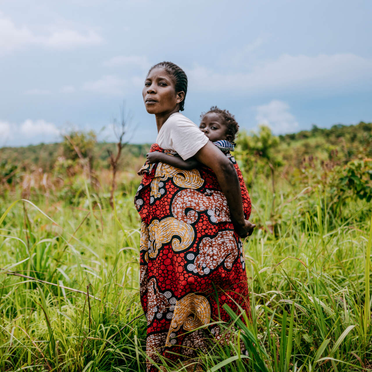 En mamma som bär sitt barn på ryggen ståendes på ett fält. 