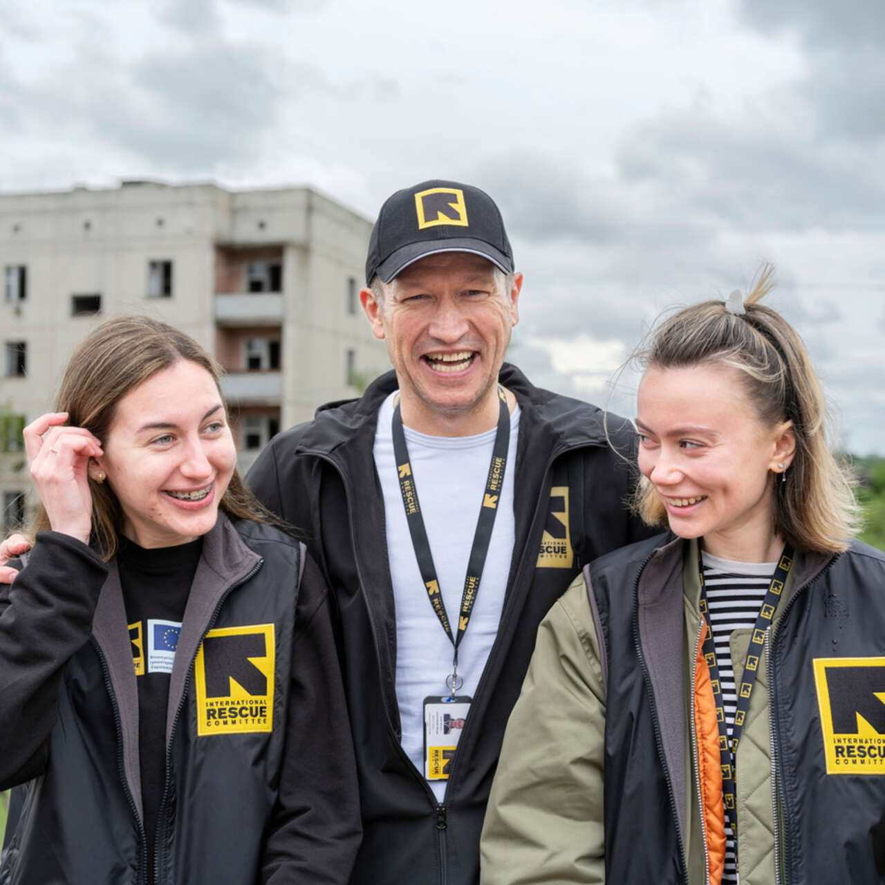 re medarbetare från RESCUE kramar om varandra utomhus i Ukraina, med en skadad byggnad i bakgrunden.