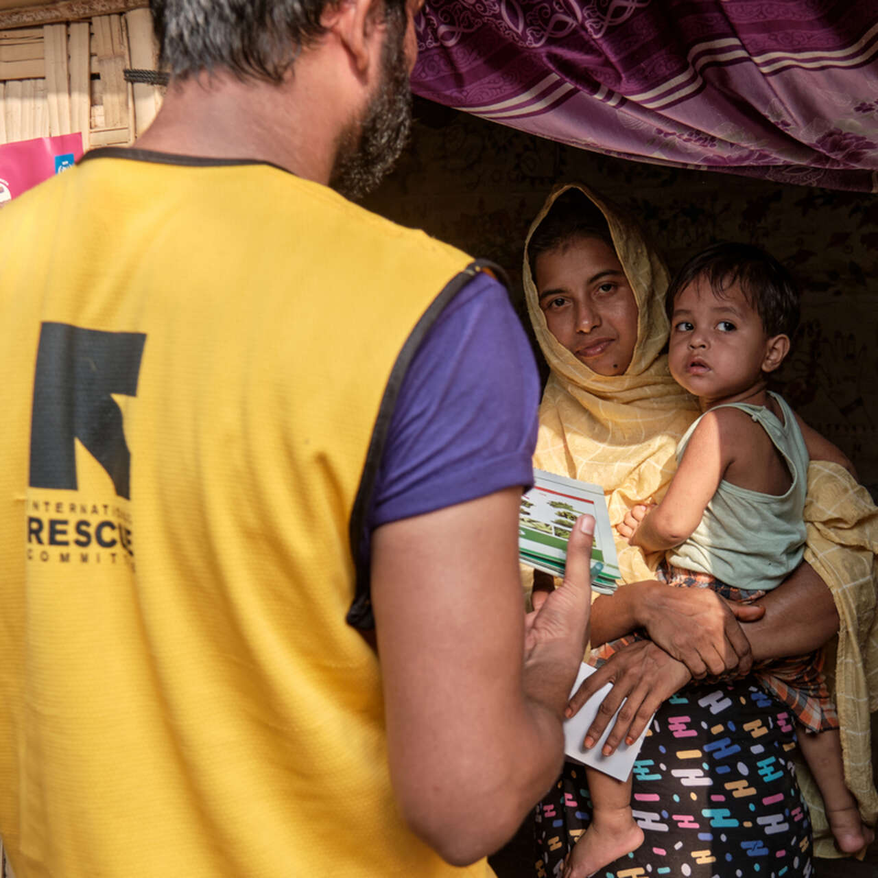 En medarbetare från RESCUE i gul väst, sedd bakifrån, ger broschyrer till en kvinna med gul sjal som håller ett litet barn i dörröppningen till ett bambuskjul i Bangladesh.
