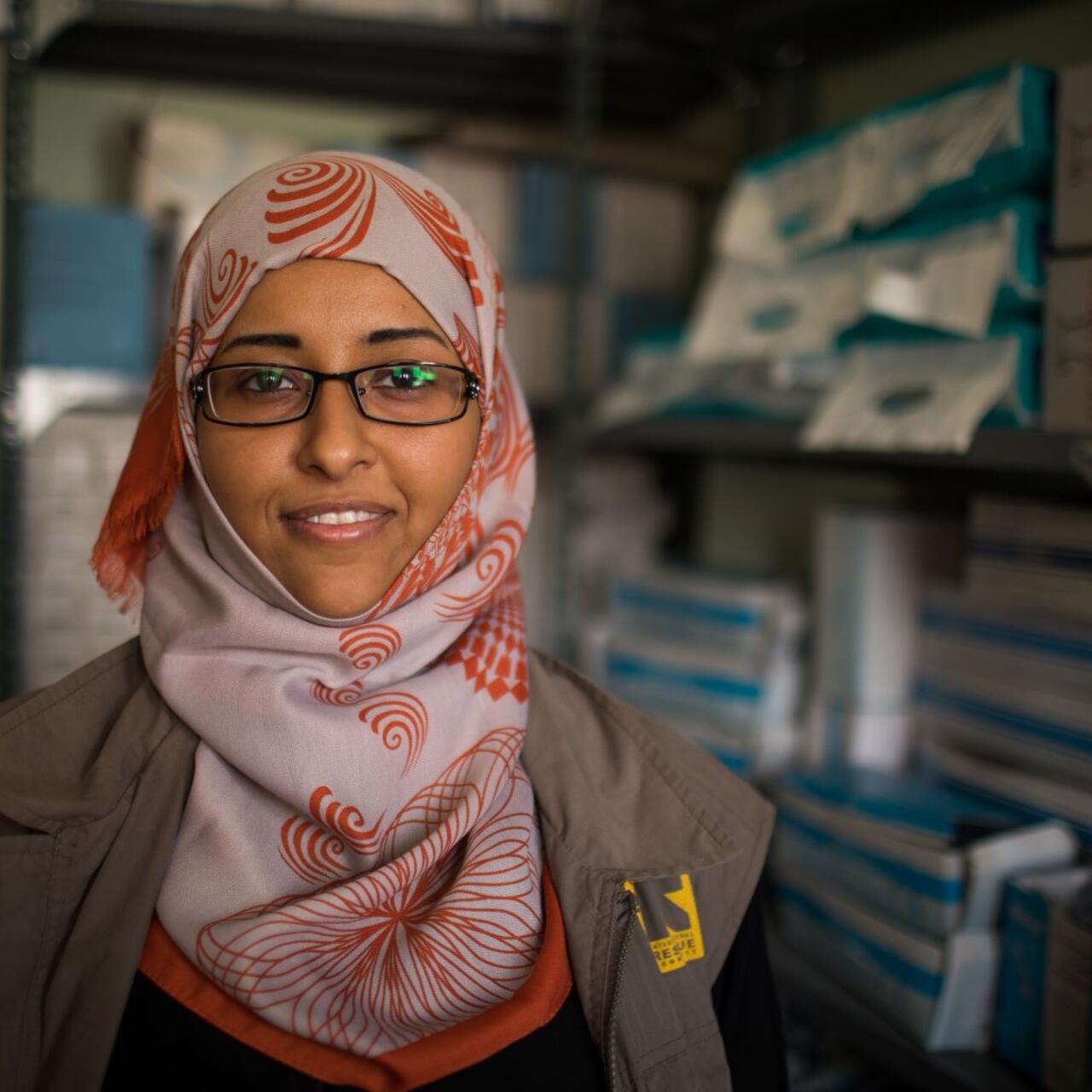 Dr. Rasha Rashed, a doctor and reproductive health manager for the IRC in Yemen, stands in a room with media supplies. She is wearing a red and white scarf and an IRC-branded vest.