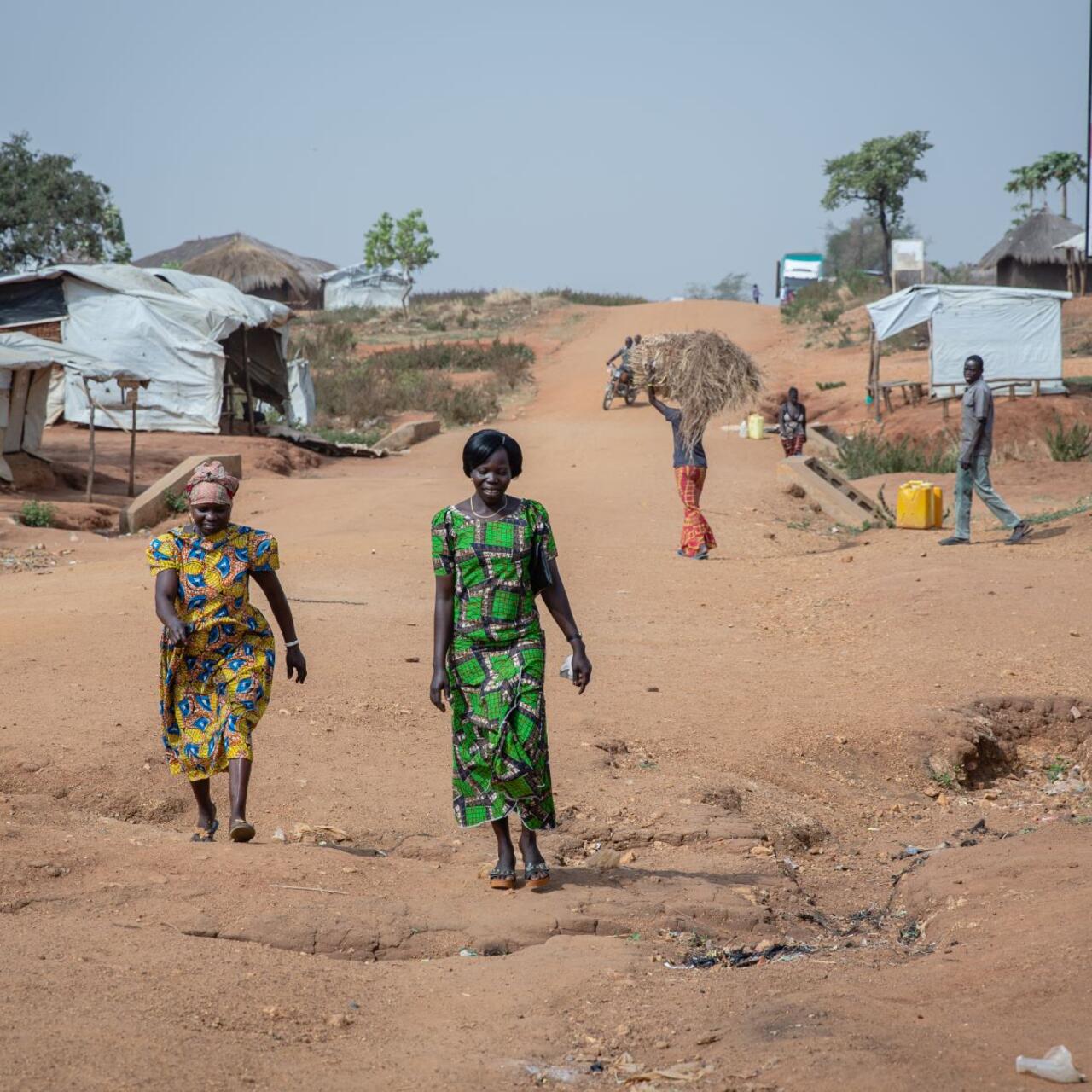 Women's rights activists Foni Grace and Loyce Tabu walk in Bidi Bidi refugeee settlement in Uganda.They are at the forefront of the photo and there are small makeshift homes and a man carrying a bale of hay behind them.