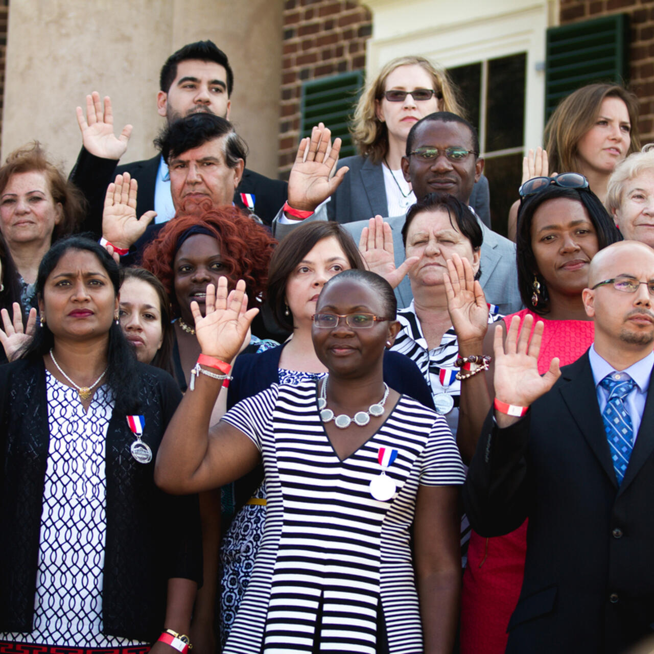 Two new citizens take their oath. They are standing in a large group of people and holding up their right hands. 