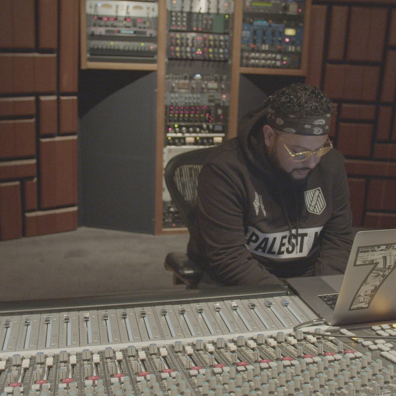 Belly, wearing a headband and a "Palestine" sweatshirt, sits at his laptop, which is perched on a mixing board in a recording studio.