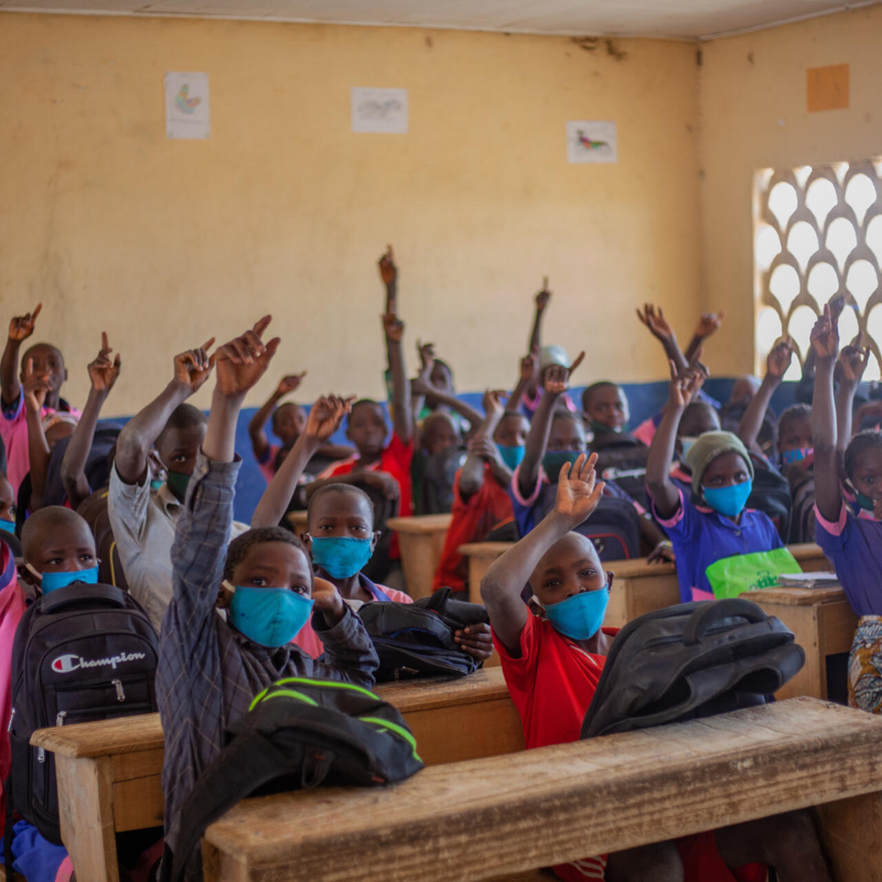 Children seated at wooden desks raise their hands in a classroom with breeze block windows in northern Cameroon.
