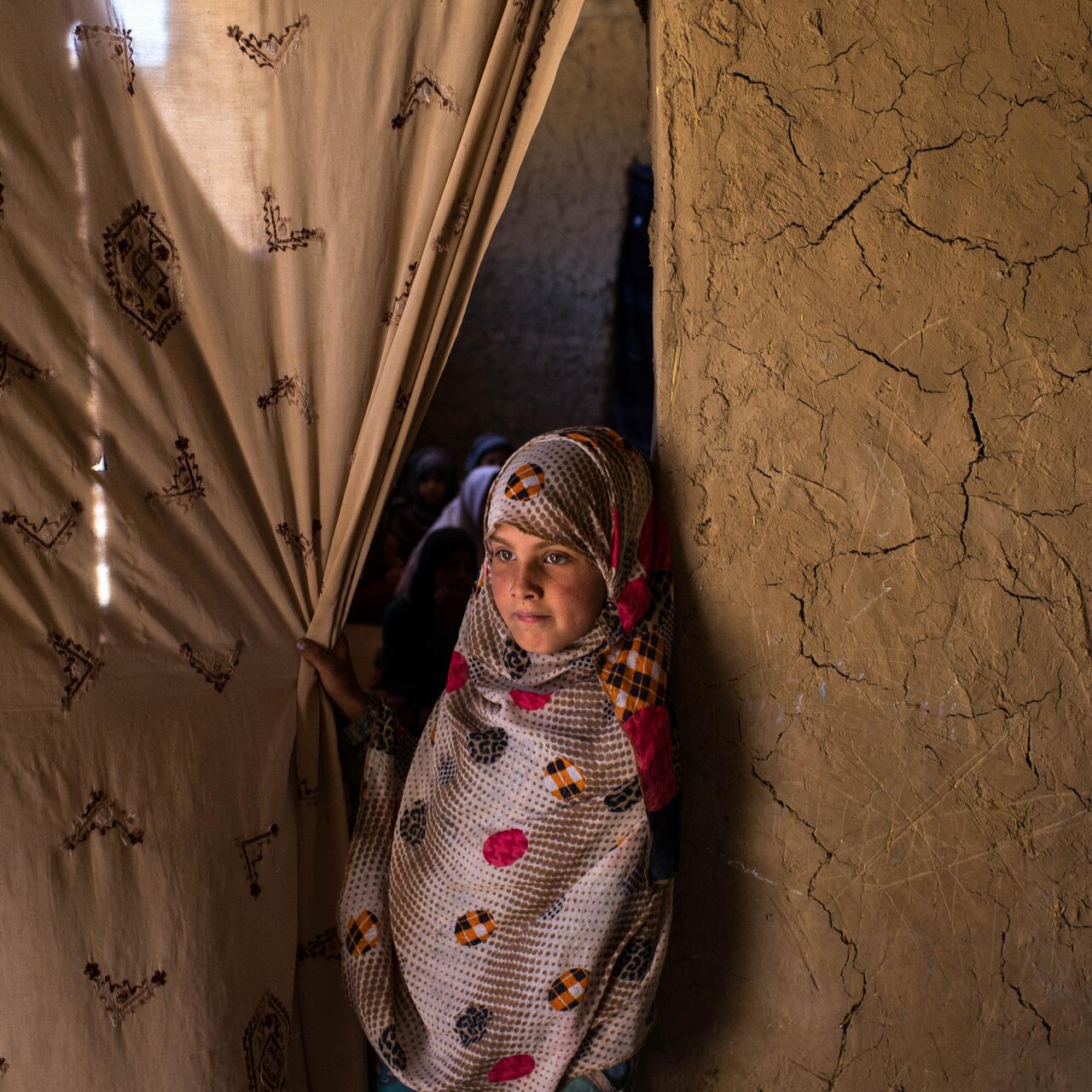 A young girl grasps a curtain hanging in a doorway in Afghanistan, looking out as she leans agains the wall.