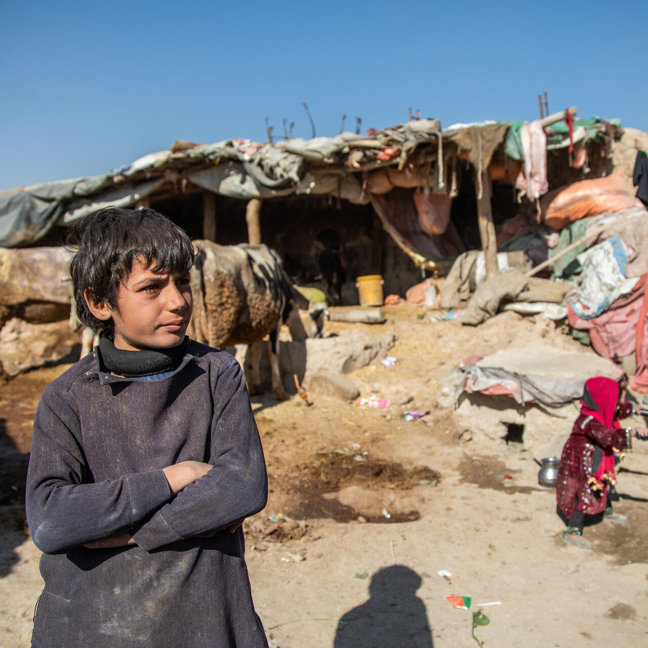 In front of a makeshift shelter, a boy stands with his arms crossed.