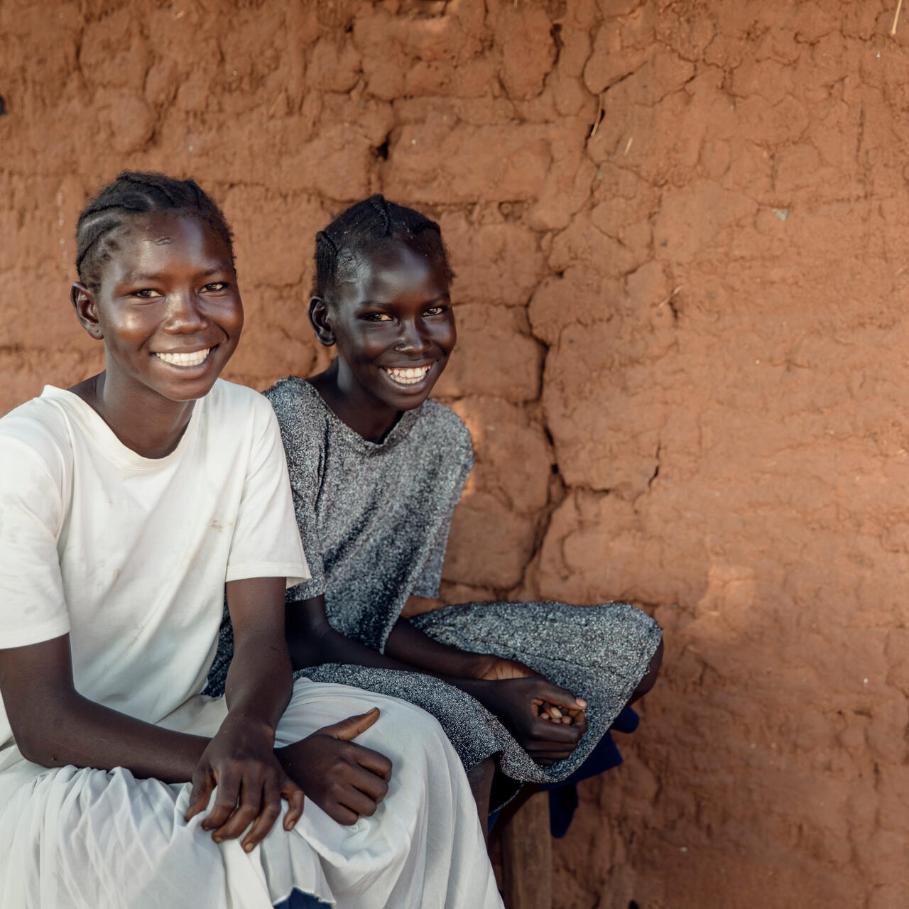 Two young girls sit on the edge of a bed and smile looking at the camera.