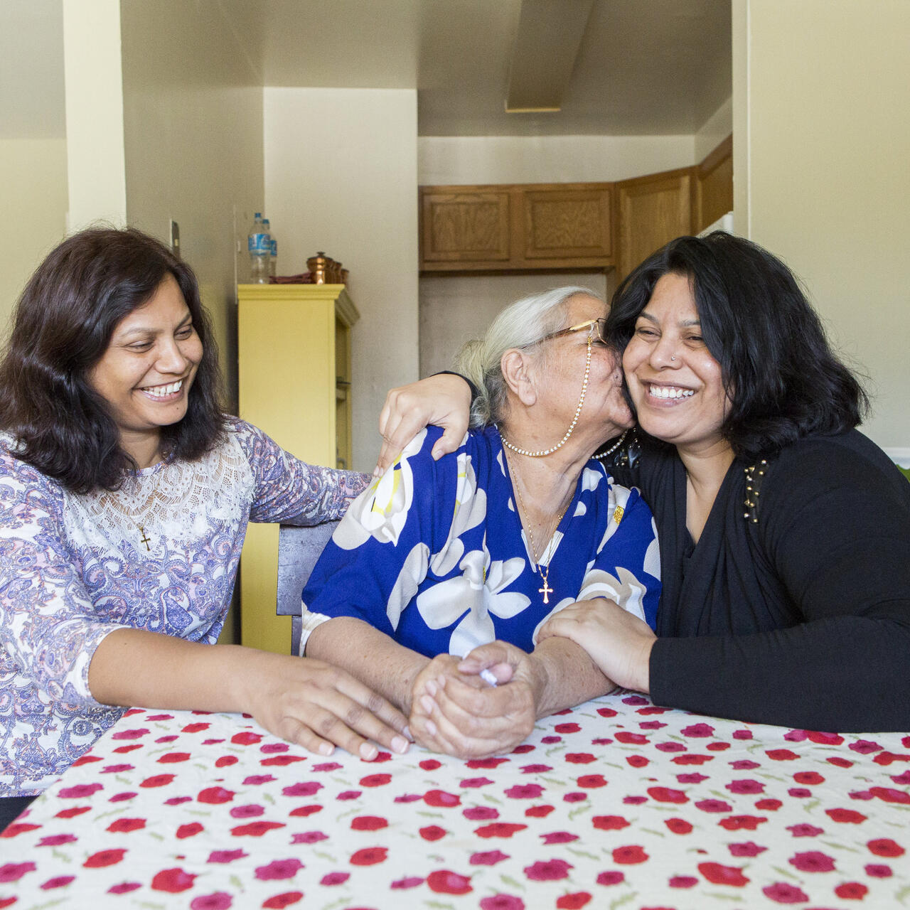 Three women, one older and two middle aged, sit at a table with a red and white table clothe. They are smiling and the older woman is kissing the woman on her left. They are all refugees. 