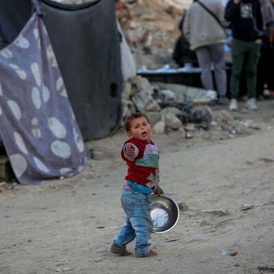 A child in a red sweater and blue jeans stands in a dusty area in Gaza City holding a metal bowl. 