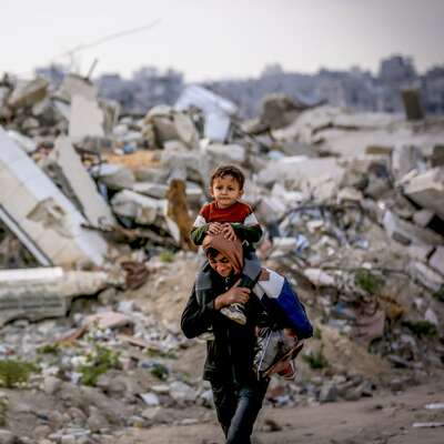 A person carries a child on their shoulders walking through rubble in the Jabalia Refugee Camp, Gaza