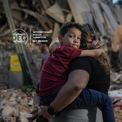 A mother holds her son as they walk past debris from the earthquake in Venezuela. Text donates the IRC as a DEC partner.
