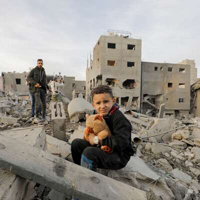 A boy clutches a toy while a man stands nearby amidst the ruins of their neighborhood in the Nuseirat refugee camp